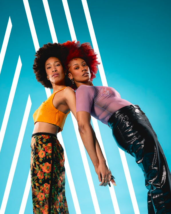 Two women pose back-to-back against a vivid blue backdrop with white diagonal light lines, showcasing bold fashion: one in an orange crochet crop top and floral pants, the other in a lilac top and shiny black pants.